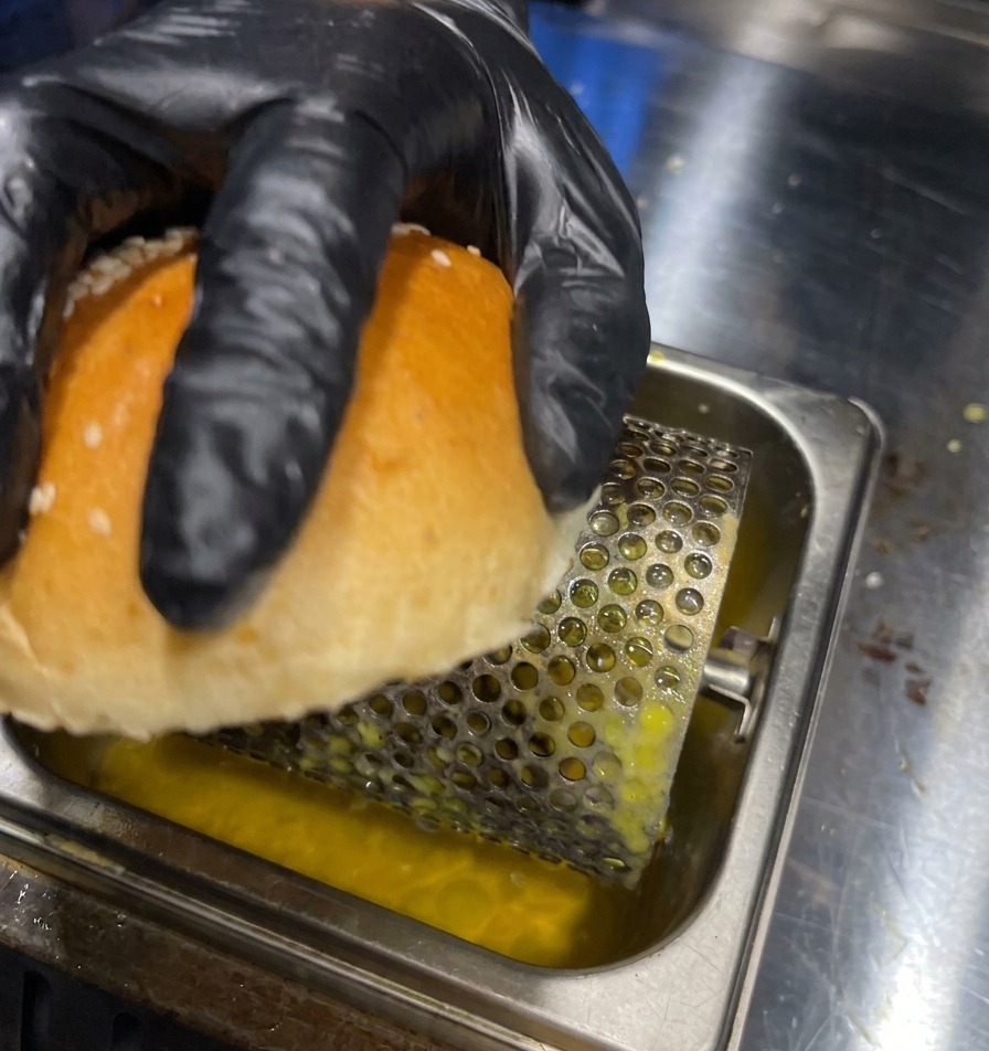 Eine Hand rollt die Innenseite eines Burgerbrötchens über eine Butterwalze in einer Metallschale.