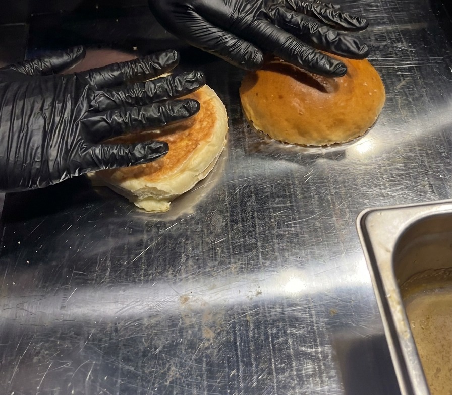 Zwei Burgerbrötchen werden mit der Hand auf eine heiße Grillplatte gedrückt, um die Innenseiten zu rösten.