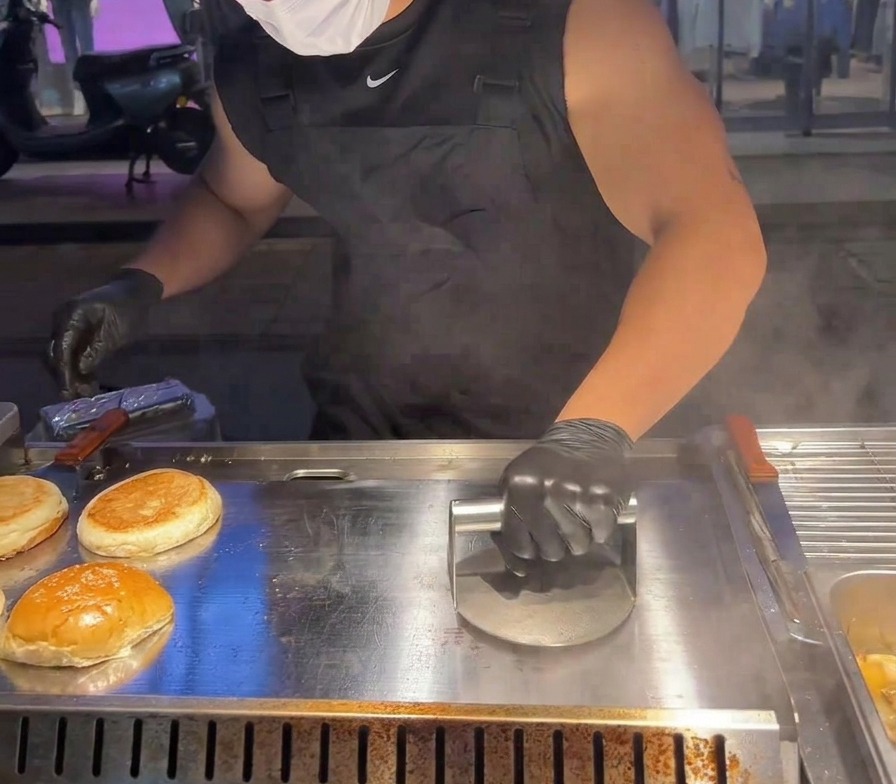 Eine schwere Metallpresse wird verwendet, um ein Fleisch-Patty auf der heißen Grillplatte flach zu drücken.
