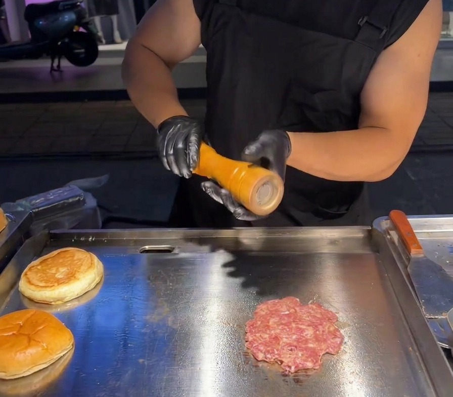 Frisches Meersalz wird aus einer Mühle über das flachgedrückte Patty auf einer Grillplatte gemahlen.