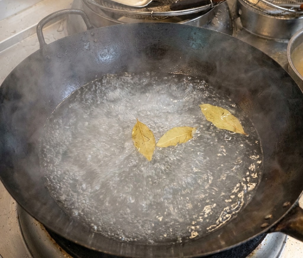 Ein großer schwarzer Topf mit kochendem Wasser, in dem drei Lorbeerblätter an der Oberfläche schwimmen.