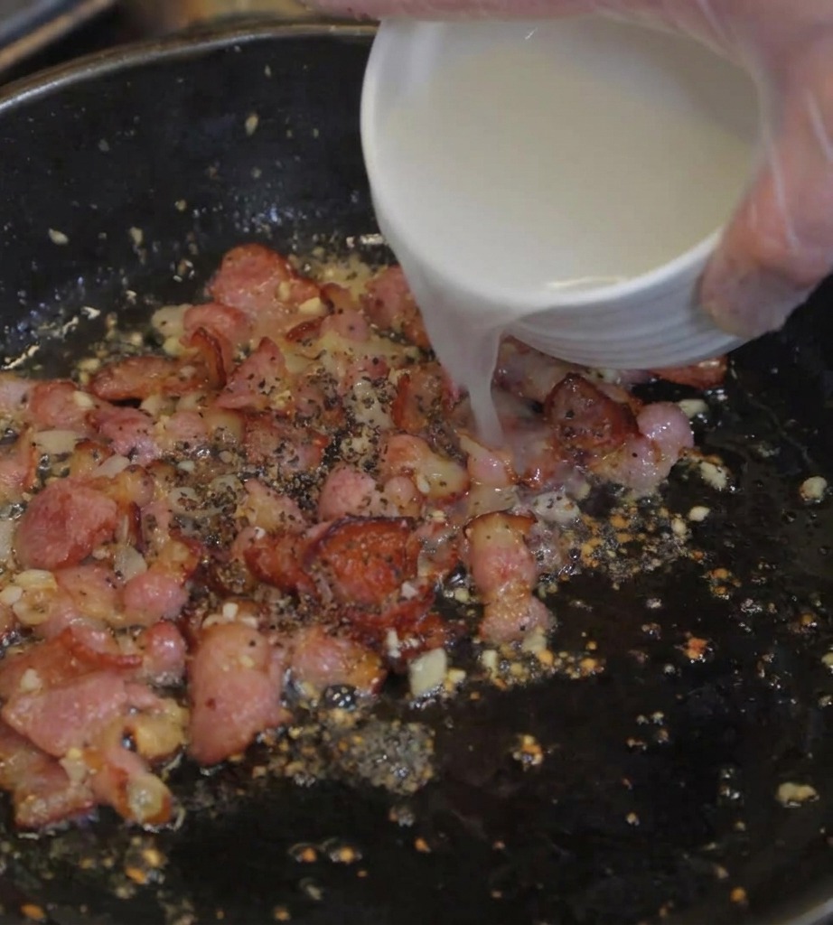 Trübes Nudelwasser wird aus einer Schüssel in die Pfanne mit Speck und Zwiebeln gegossen.