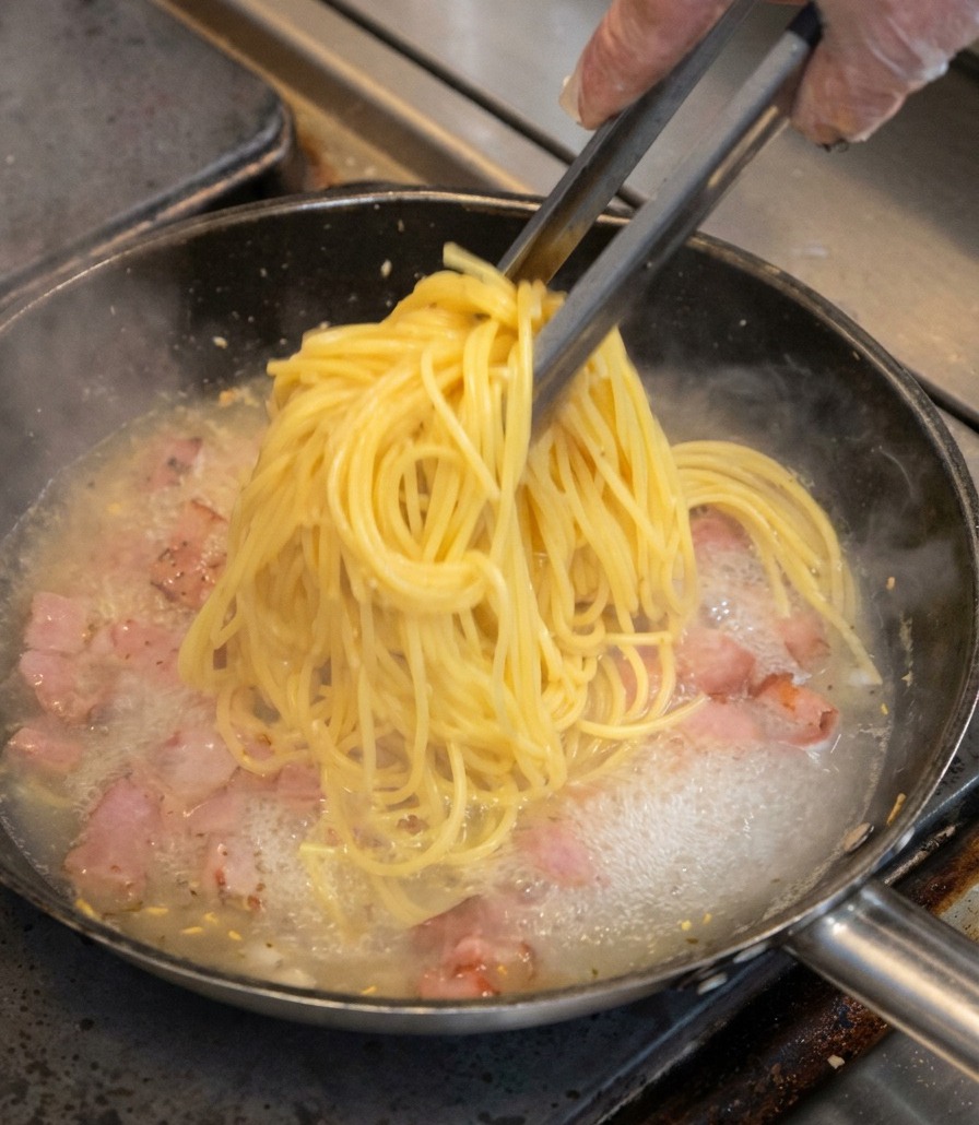 Ein Bündel Spaghetti wird mit einer Küchenzange in die Pfanne zum Speck gehoben.
