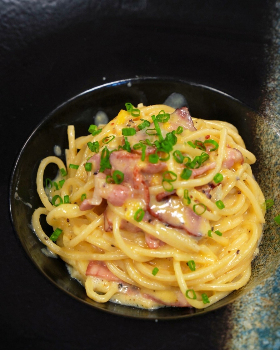 Eine fertige Portion Carbonara wird in einer schwarzen Schüssel mit frischem Schnittlauch bestreut.