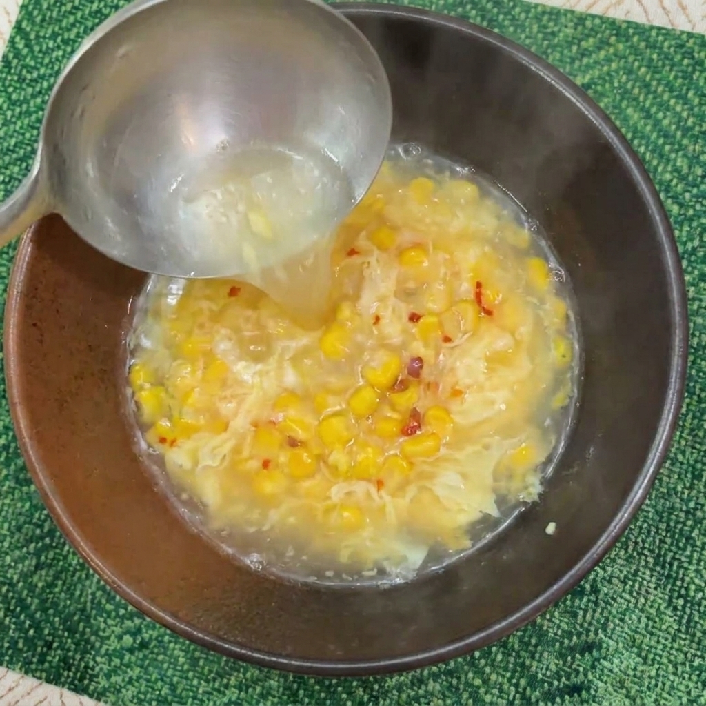 Eine Schöpfkelle gießt dampfend heißes Wasser in eine Schüssel mit Mais und der Eimischung.