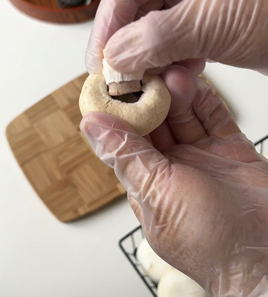 Eine Person mit Kunststoffhandschuhen entfernt den Stiel eines Champignons über einem Holzbrett.