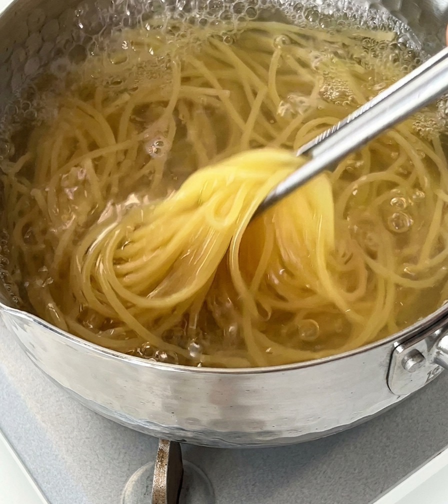 Nahaufnahme von Spaghetti, die mit einer Metallzange aus kochendem Wasser gehoben werden.