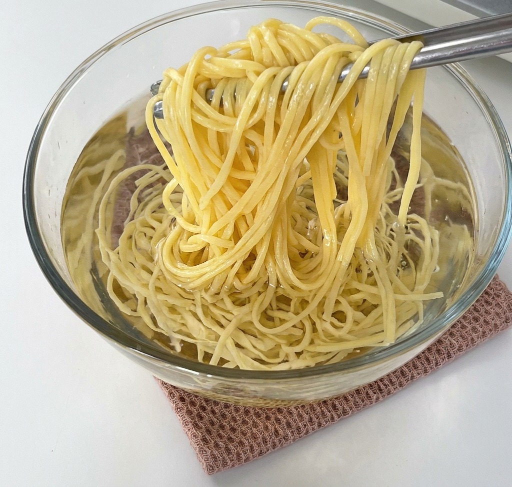 Eine Zange hebt gekochte Spaghetti aus einer Glasschüssel mit kaltem Wasser.