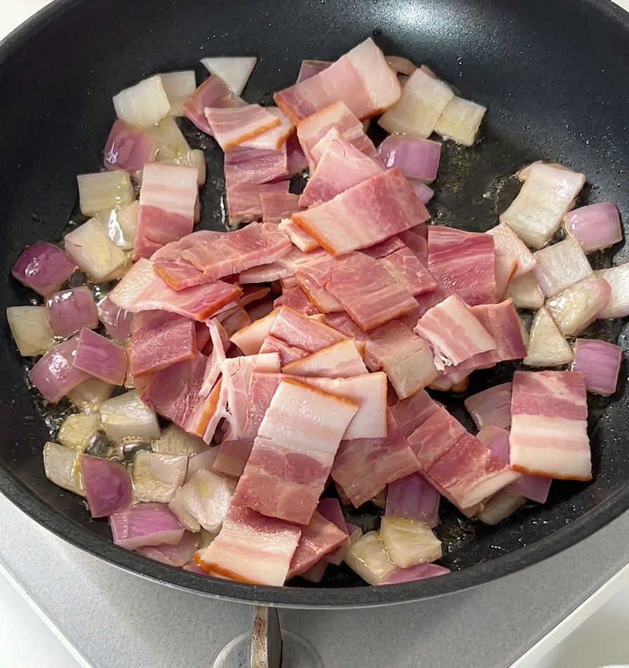 Rohe Speckstücke liegen auf gedünsteten Zwiebeln in einer Pfanne.
