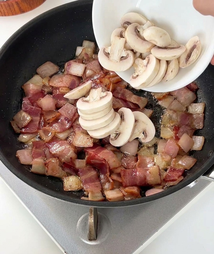 Eine Person gibt geschnittene Champignons in die Pfanne zu Speck und Zwiebeln.