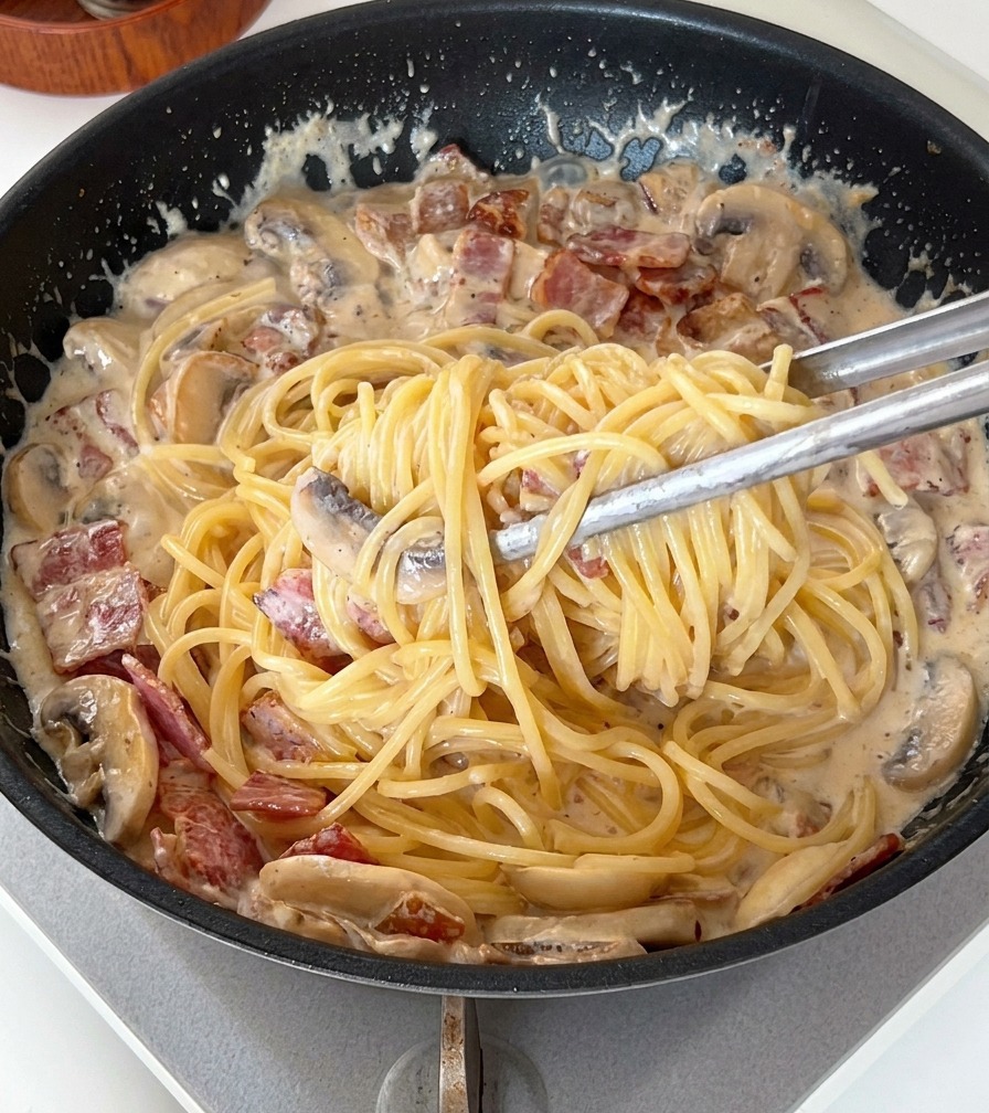 Eine Zange mischt die Spaghetti in der cremigen Sauce mit Speck und Pilzen.