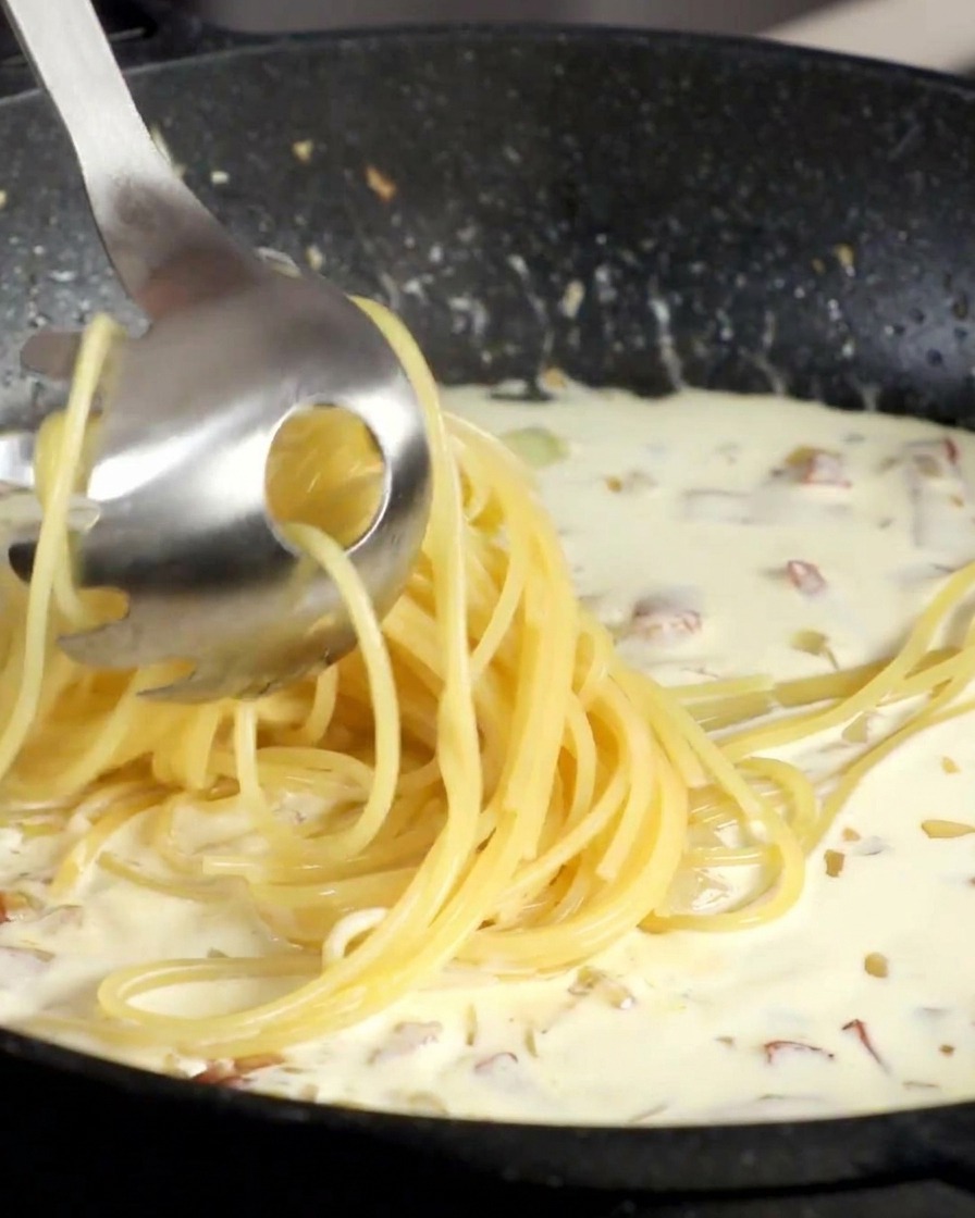 Spaghetti werden mit einer Nudelzange direkt in die Sahnesauce gehoben.