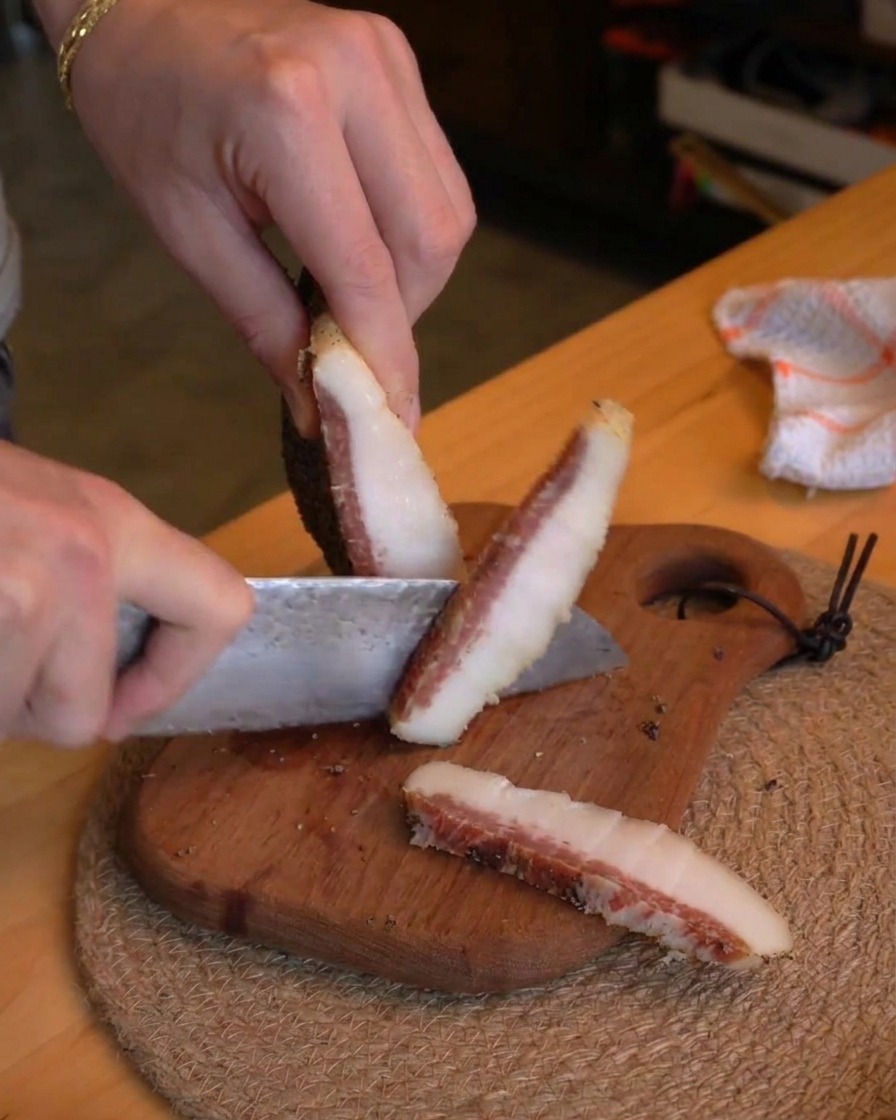 Eine Person schneidet mit einem Kochmesser dicke Streifen Guanciale in kleine Würfel auf einem rustikalen Holzbrett.