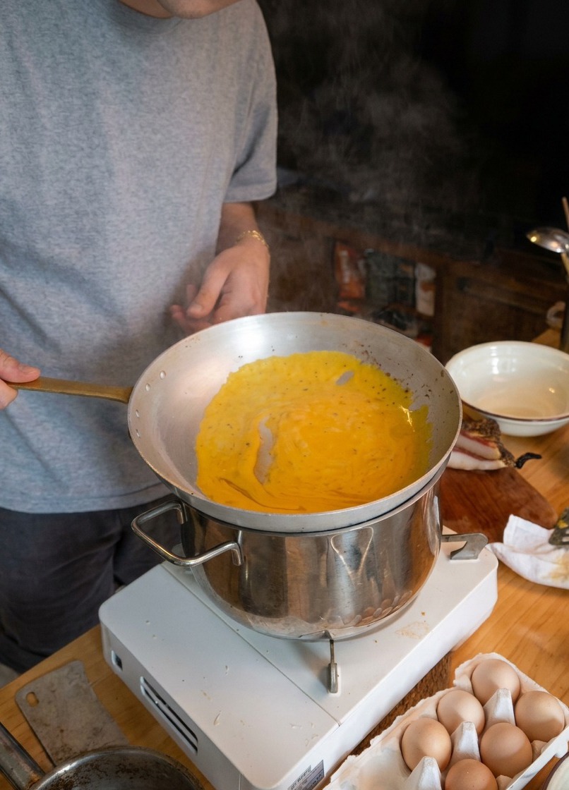 Ein Koch schwenkt Spaghetti in einer Metallpfanne über dem dampfenden Topf, um die Sauce zu binden.