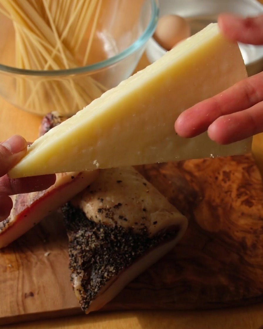 Nahaufnahme einer Hand, die ein Stück Pecorino Romano über zwei Scheiben Guanciale auf einem Holzbrett hält.