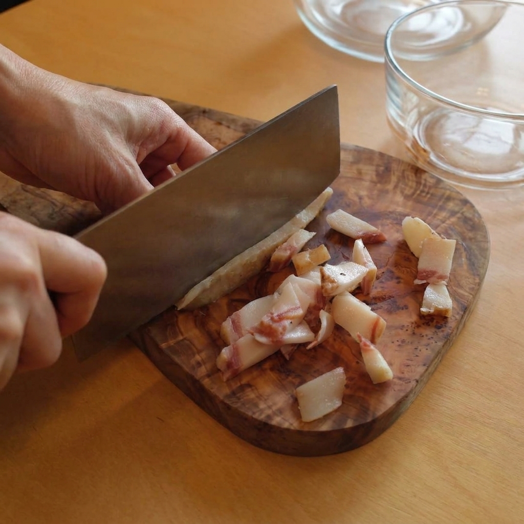 Ein Küchenmesser schneidet Streifen von fettiger Guanciale auf einem rustikalen Holzbrett.