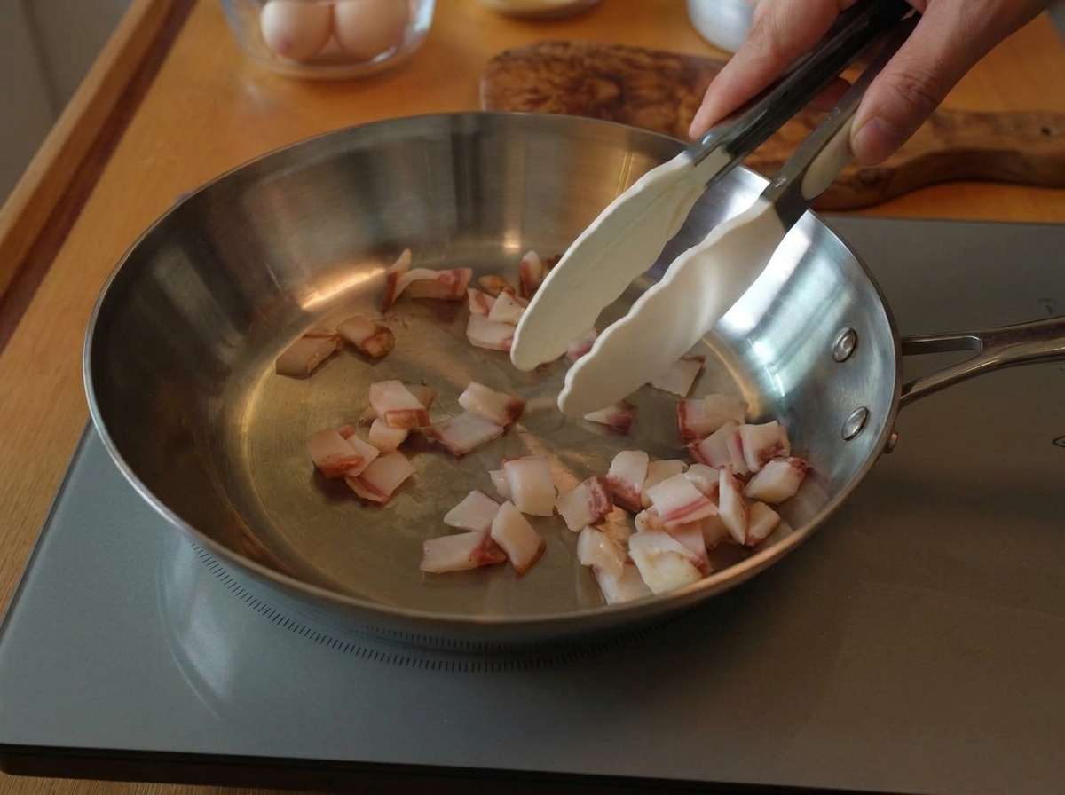 Eine Zange rührt Guanciale-Stücke in einer Edelstahlpfanne auf einem Induktionsherd.