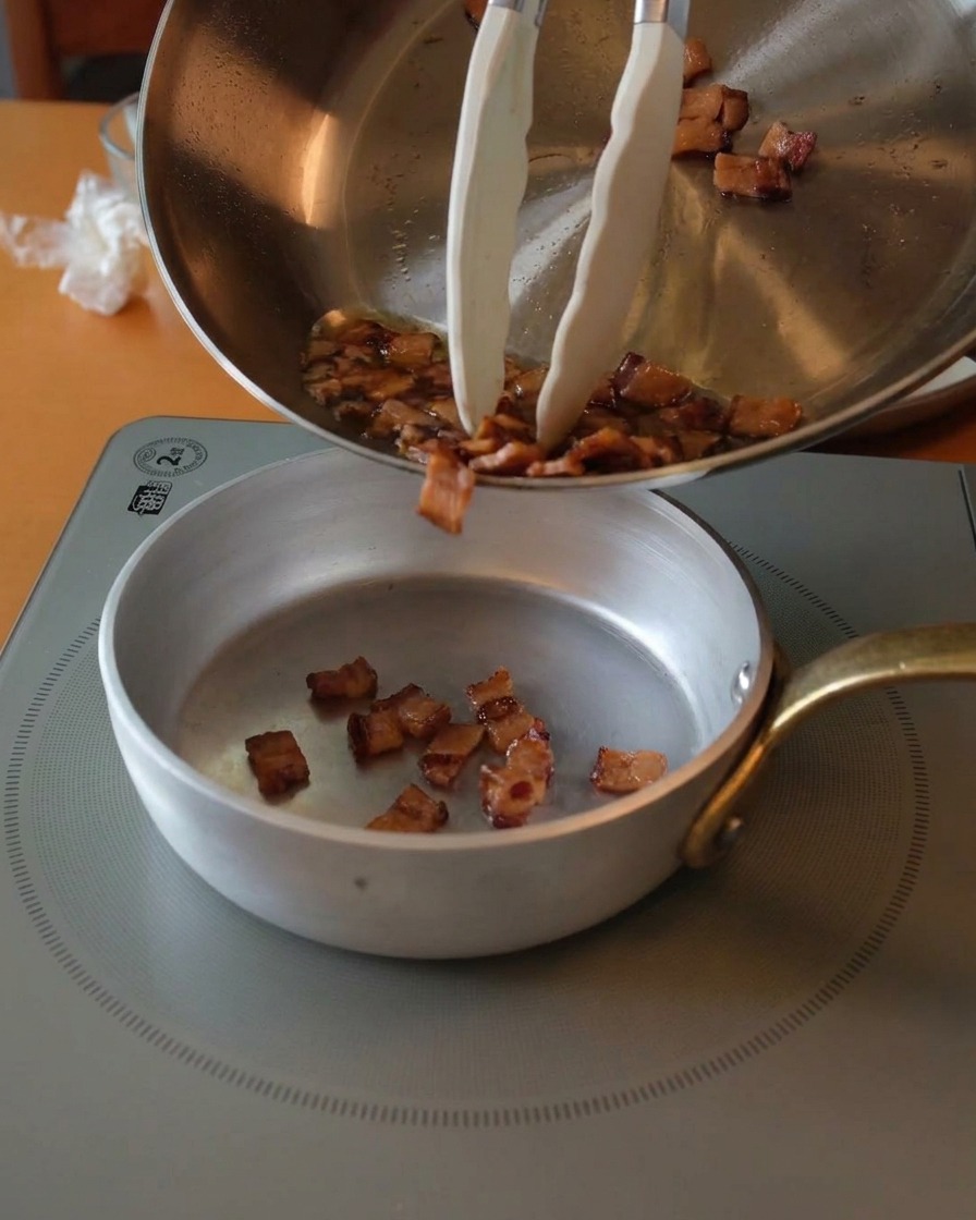 Knusprige goldbraune Guanciale-Stücke werden mit einer Zange aus einer Pfanne in eine kleine Silberschale gefüllt.