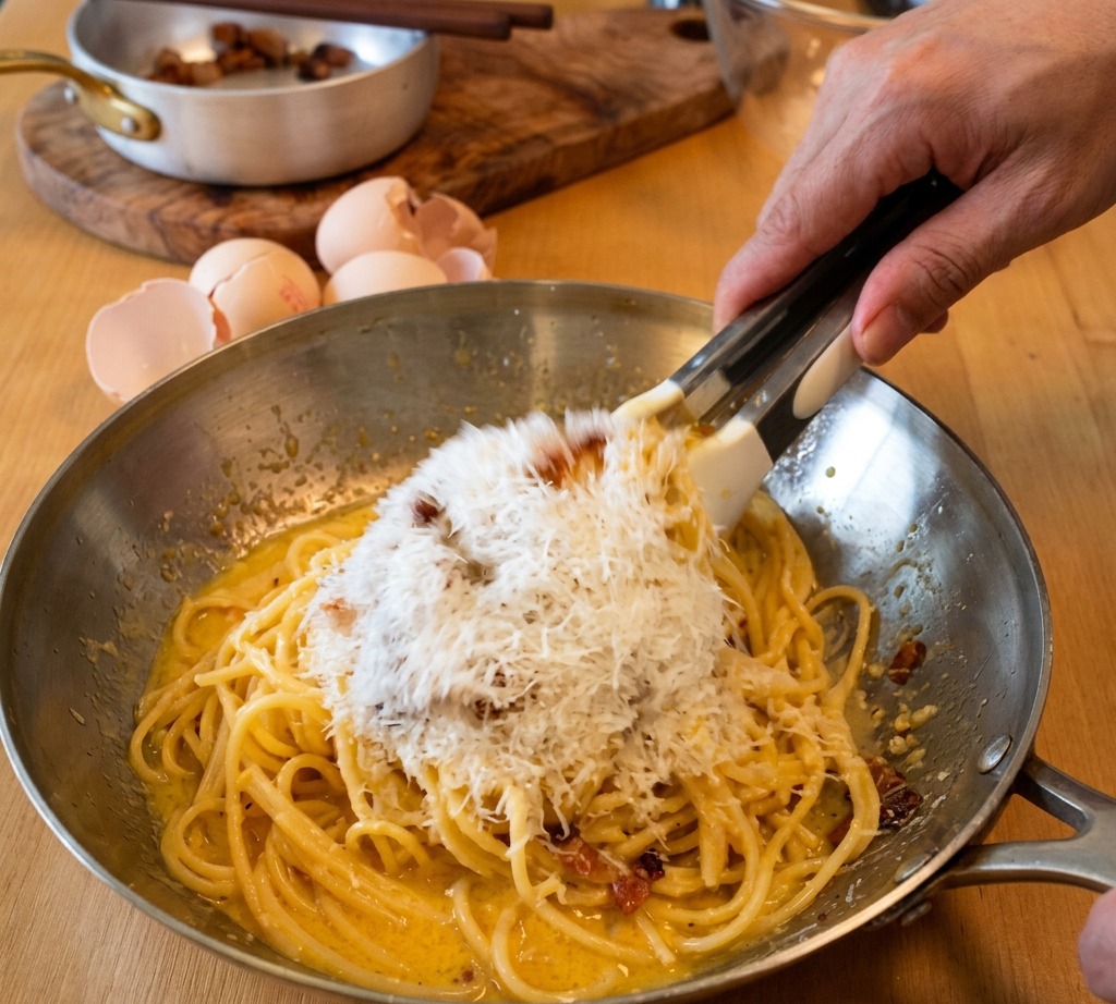 Spaghetti werden in einer Pfanne geschwenkt, während fein geriebener Pecorino Romano zur Eigelbmischung gegeben wird.