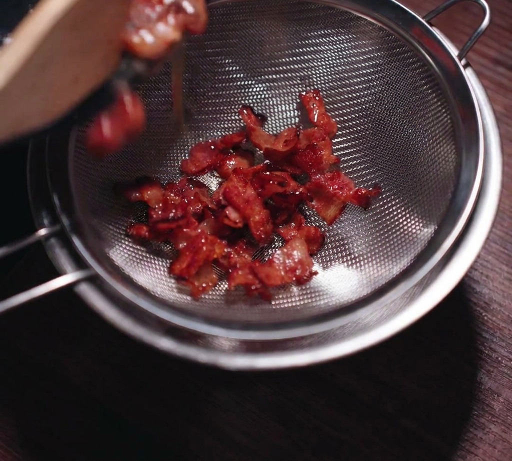 Knusprig gebratener Speck wird durch ein Edelstahlsieb gegossen, um das flüssige Fett aufzufangen.