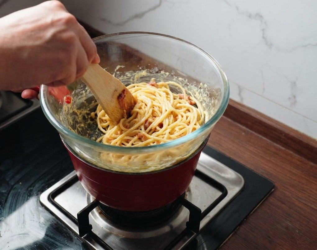 Pasta und Speck werden mit einem Spatel in der goldenen Sauce vermengt, während die Schüssel über Dampf steht.