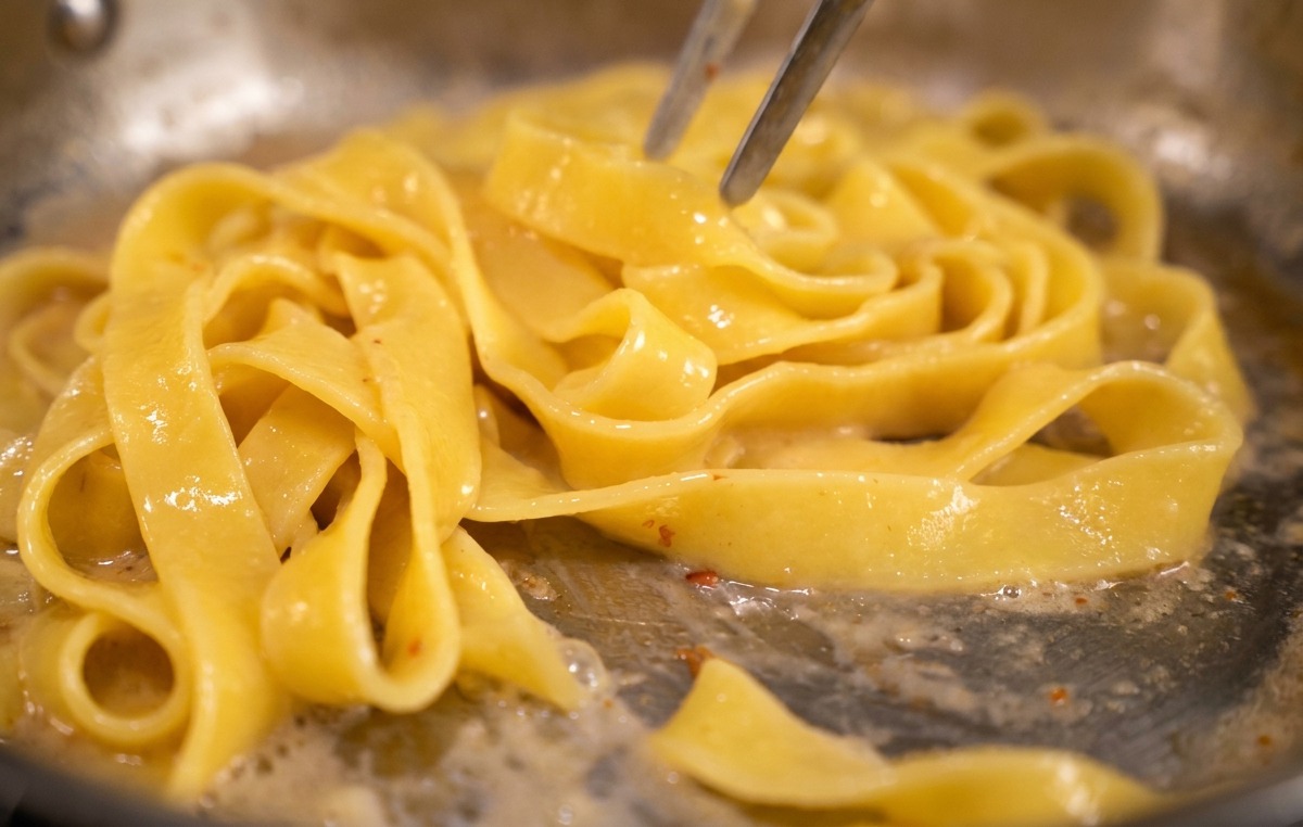 Pasta wird mit einer Zange in einer Pfanne geschwenkt, um die gelbe Eigelbsauce zu binden.
