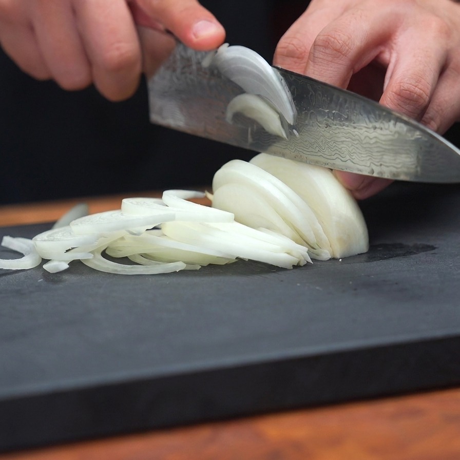 Nahaufnahme einer Person, die mit einem scharfen Messer gleichmäßige, dünne Scheiben einer weißen Zwiebel auf einem grauen Schneidebrett schneidet.
