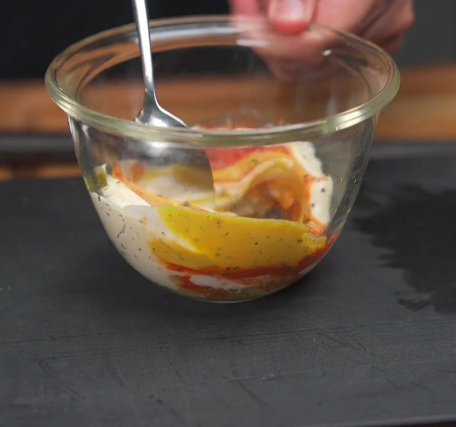 Ein Löffel rührt roten Ketchup, gelben Senf und weiße Mayonnaise in einer klaren Glasschüssel zusammen.