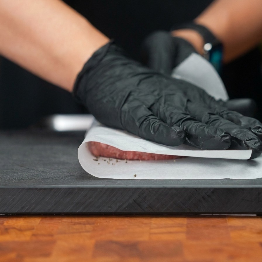 Hände mit Handschuhen drücken durch eine Schicht Backpapier auf ein Fleisch-Patty, um es zu einer breiten, dünnen Scheibe zu flachen.
