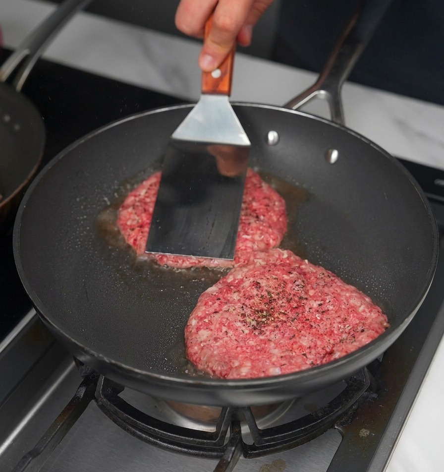 Ein Metall-Pfannenwender drückt zwei rohe Rinder-Patties in eine schwarze Pfanne, um eine Kruste zu erzeugen.