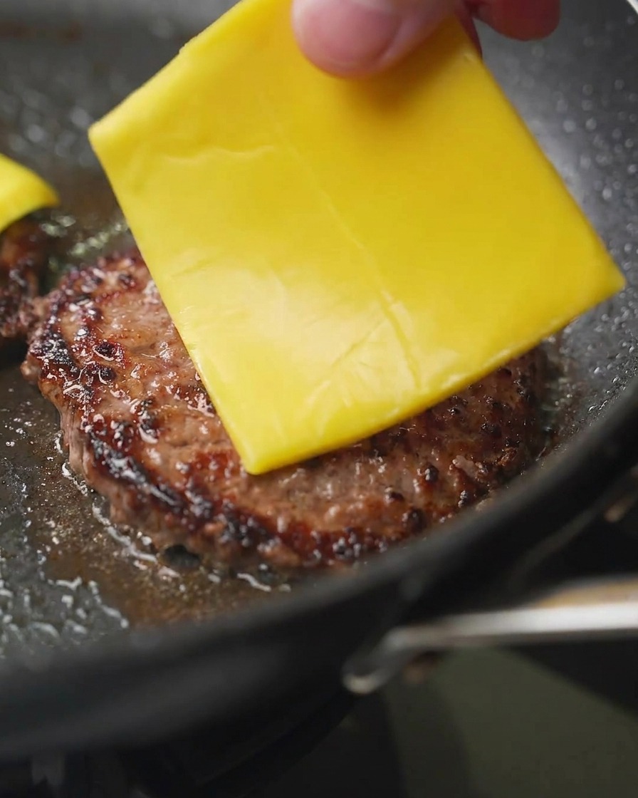 Nahaufnahme einer Hand, die eine Scheibe gelben Cheddar-Käse auf ein gebräuntes Rinder-Patty in der Pfanne legt.