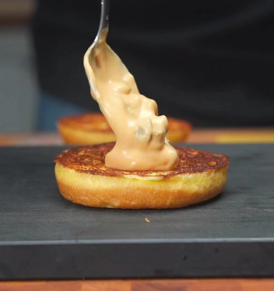 Ein Löffel verstreicht eine cremige, orangefarbene Gurkensauce auf der getoasteten Fläche eines Burgerbrötchens.