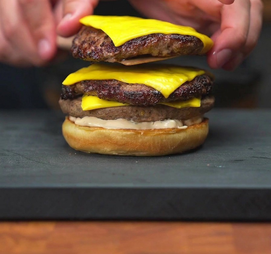 Eine Hand setzt zwei Cheeseburger-Patties mit geschmolzenem Käse auf eine bestrichene Brötchenhälfte.