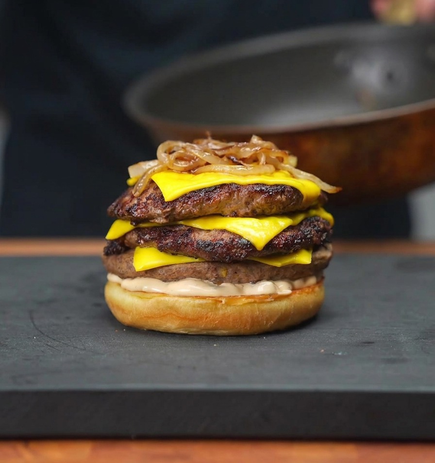Karamellisierte Zwiebeln werden mit Stäbchen auf einen Stapel Cheeseburger-Patties in einem Brötchen gelegt.