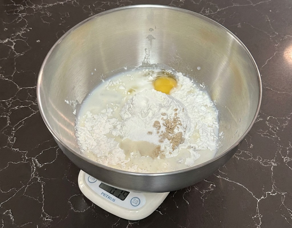 Eine Edelstahlschüssel auf einer Digitalwaage mit Mehl, Milch, einem Ei und Hefe.