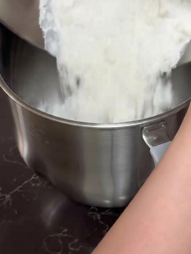 Hände gießen eine Mischung aus Mehl und Milch aus einer Edelstahlschüssel in die Schüssel einer Küchenmaschine.