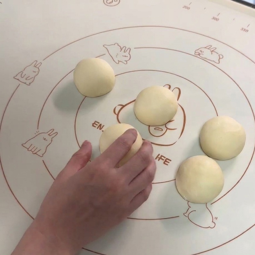 Sechs glatte, runde Teigkugeln liegen auf einer Silikonbackmatte, während eine Hand eine Portion rund wirkt.