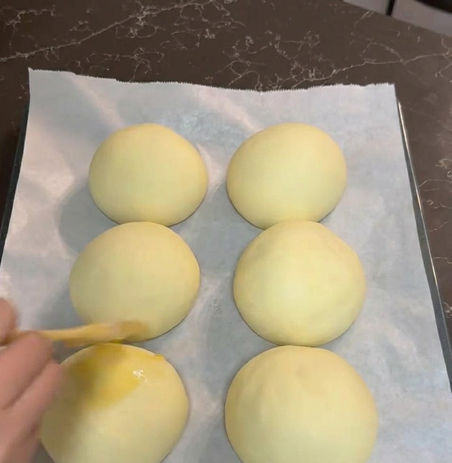 Eine Hand bestreicht sechs aufgegangene Teigkugeln mit einem Backpinsel und Eigelb auf einem Backblech.