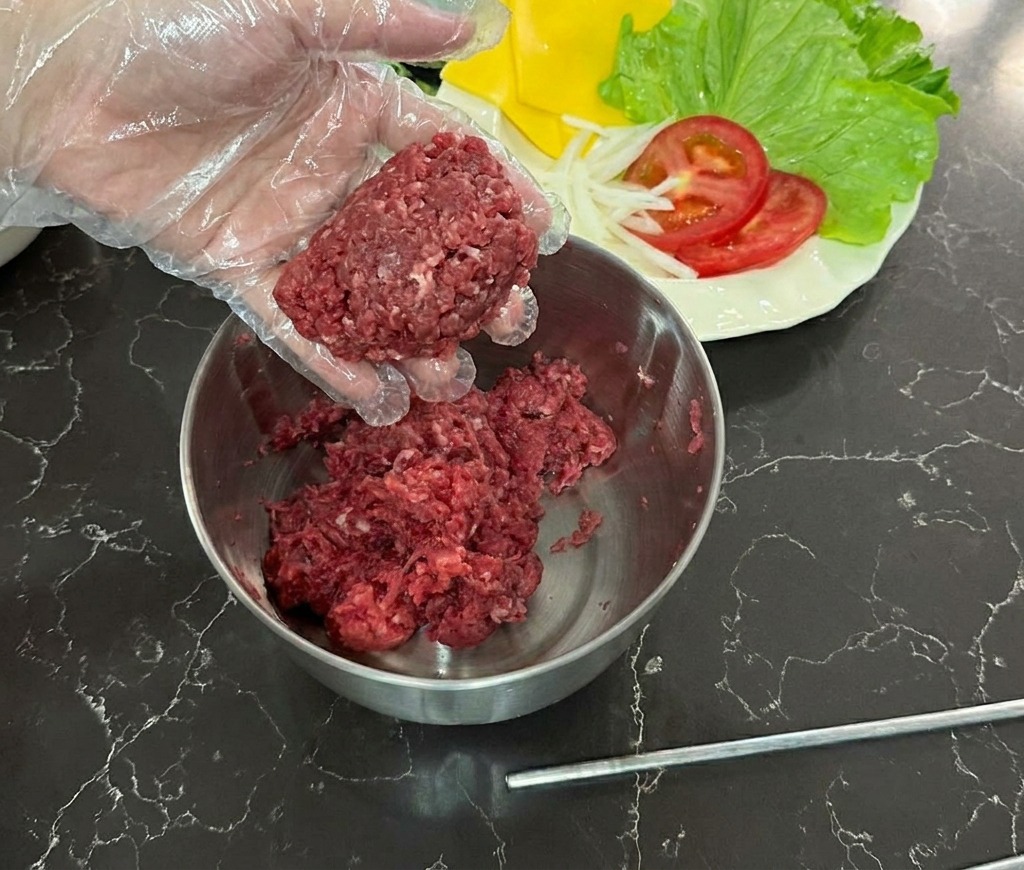 Eine Hand in einem Klarsichthandschuh hält eine Kugel aus rohem Rinderhack über eine Edelstahlschüssel.