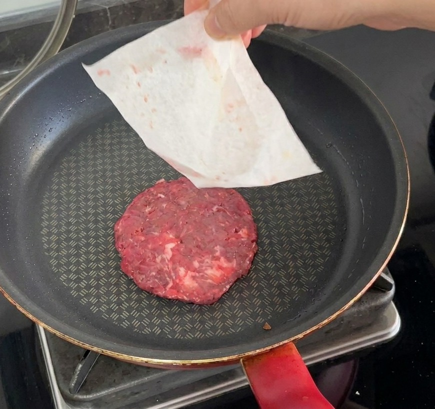 Ein rohes Patty wird in eine heiße Pfanne gelegt, während das Backpapier von Hand abgezogen wird.