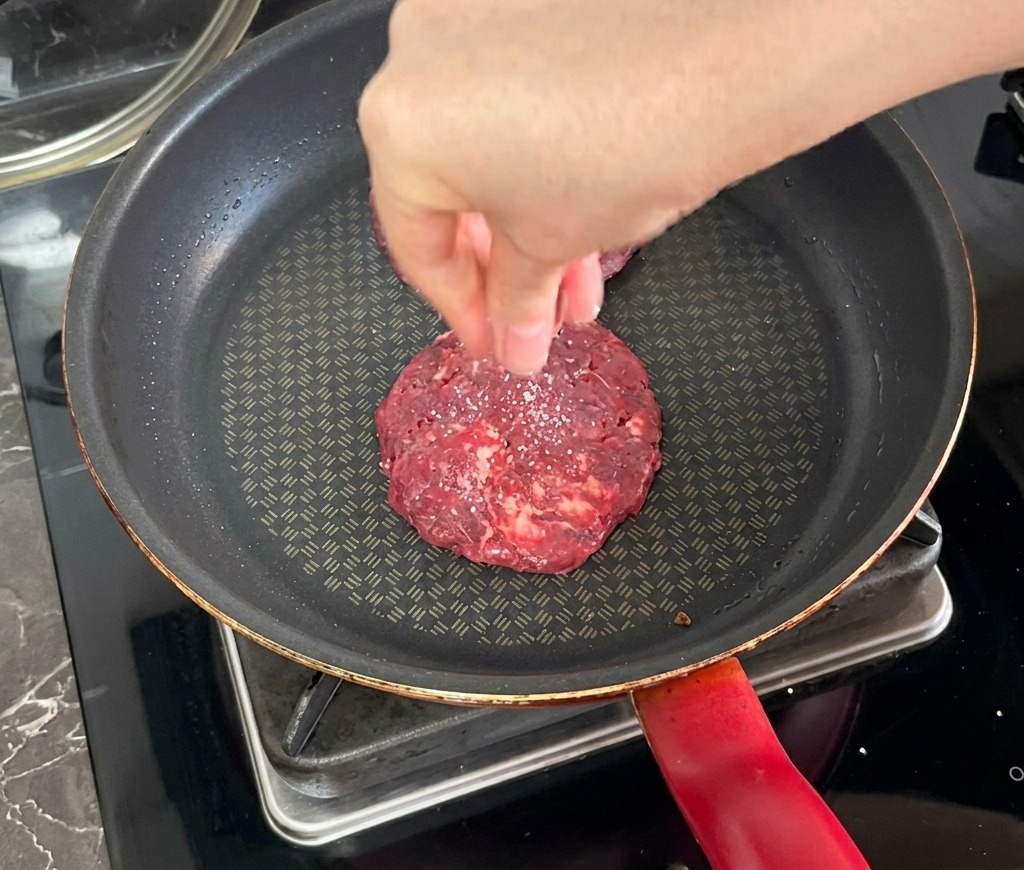 Eine Hand streut Salz über ein Rinder-Patty, das in einer dunklen Pfanne brät.