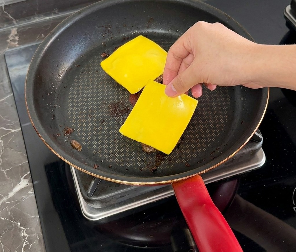 Eine Hand legt eine Scheibe gelben Käse auf ein gegartes Fleisch-Patty in einer Pfanne.