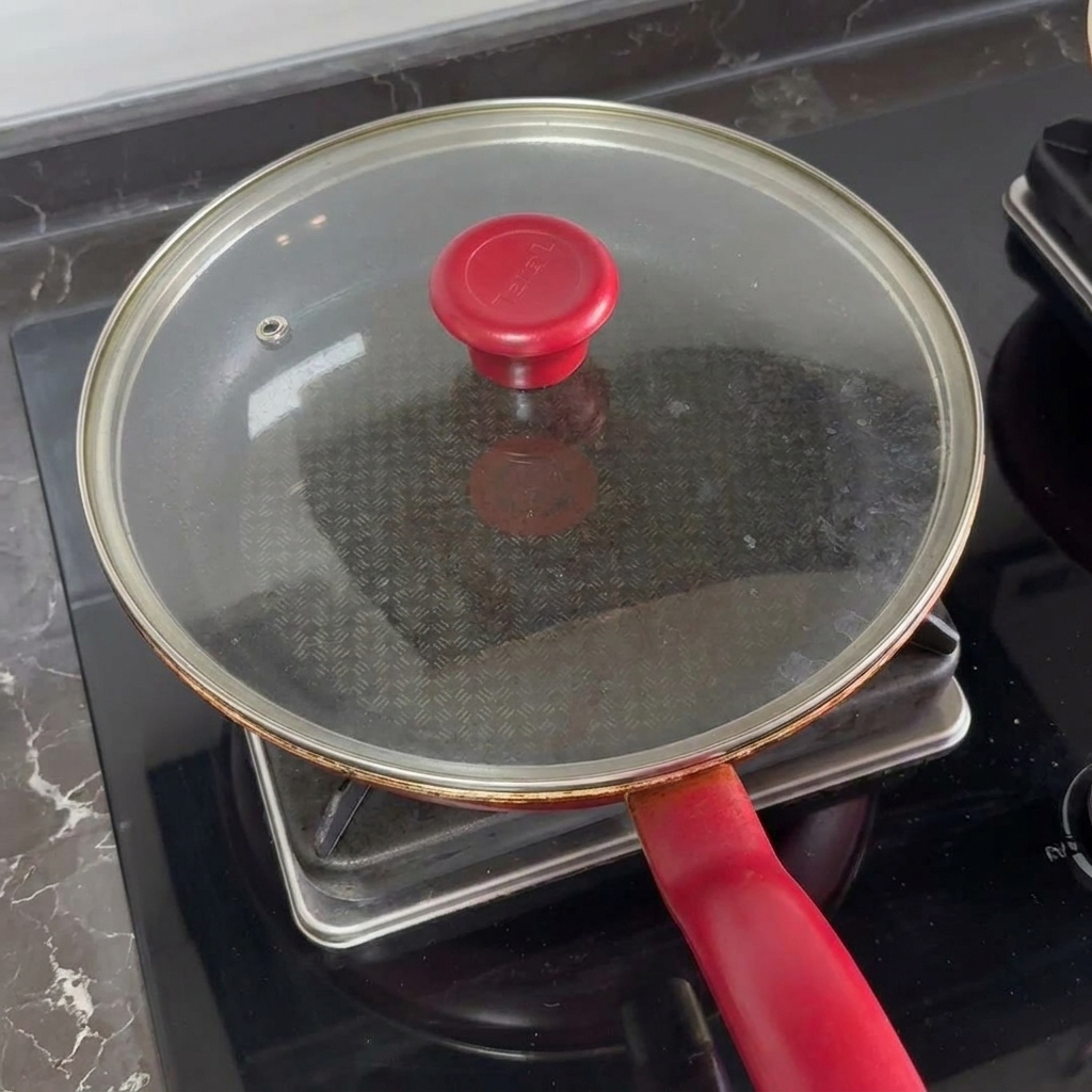 Ein Glasdeckel mit rotem Griff bedeckt die Pfanne auf dem Herd, um den Käse auf den Patties zu schmelzen.