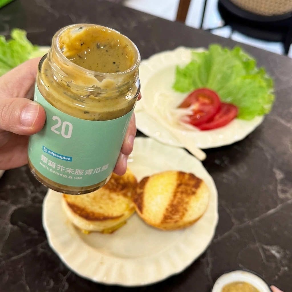 Eine Hand hält ein offenes Glas mit Senf-Gurken-Sauce über geröstete Burgerbrötchen.