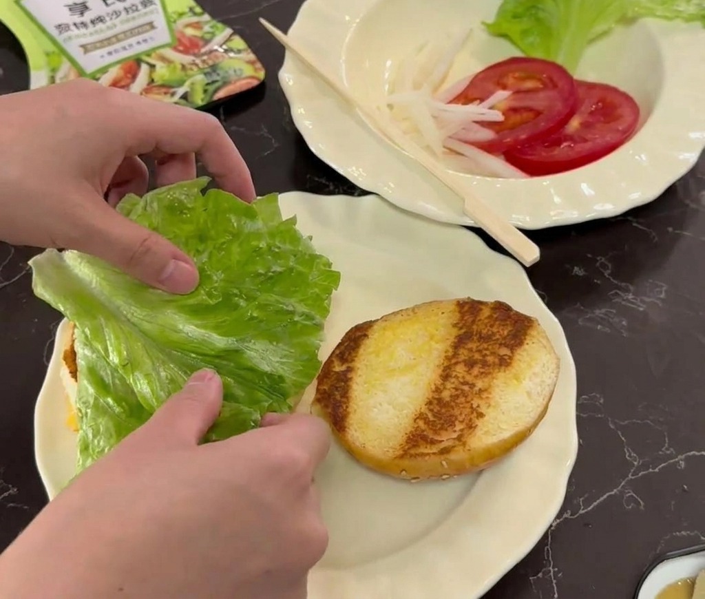 Hände legen ein frisches grünes Salatblatt auf ein geröstetes Bun auf einem weißen Teller.