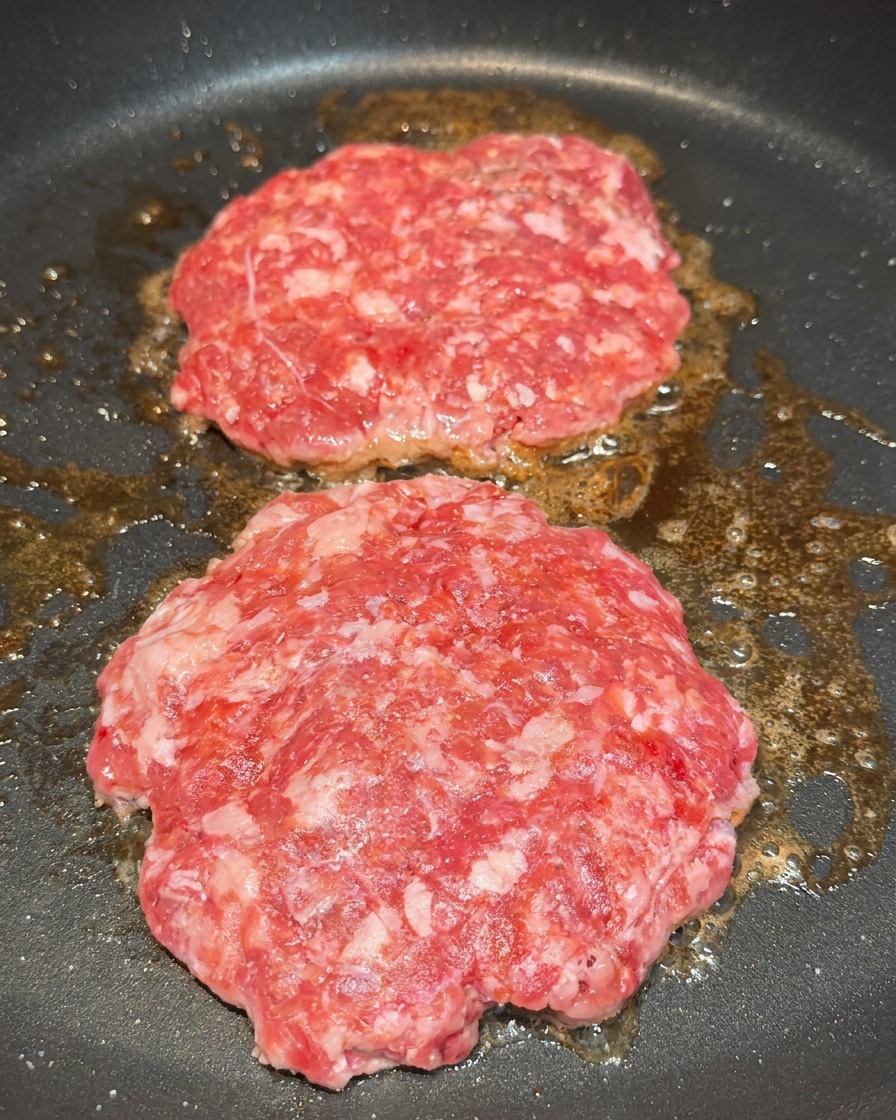 Zwei rohe Patties in einer Pfanne werden mit sichtbaren Salz- und Pfefferkörnern gewürzt.