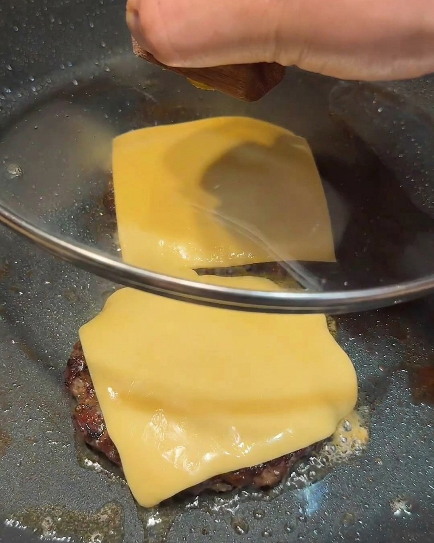 Ein Glasdeckel deckt eine Pfanne ab, in der zwei Cheeseburger-Patties braten.