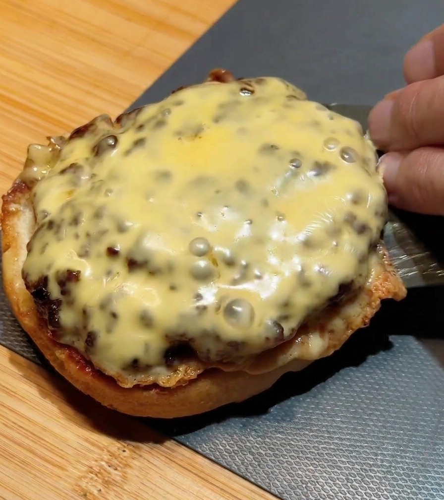 Ein Metallspatel stapelt ein zweites Cheeseburger-Patty auf das erste, das bereits auf dem unteren Bun liegt.