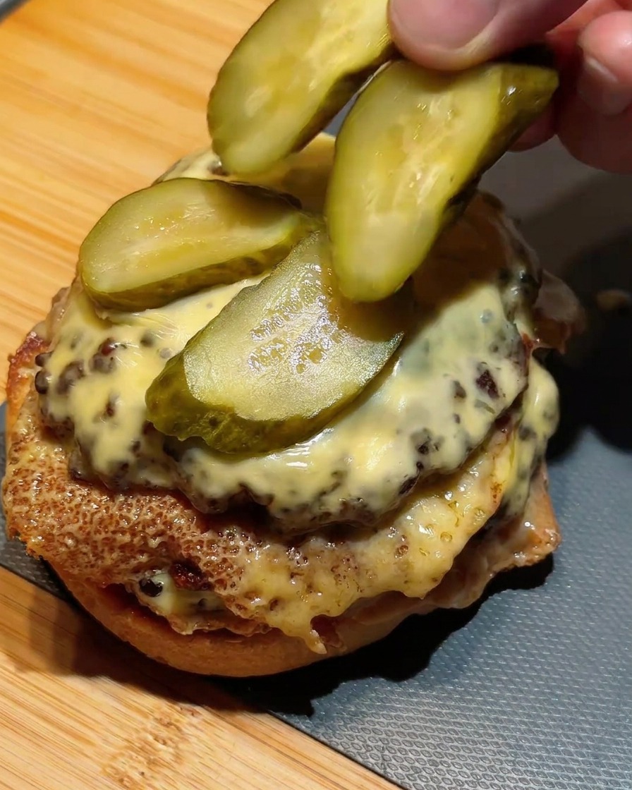 Finger legen grüne Gewürzgurkenscheiben auf den gestapelten Double Cheeseburger.
