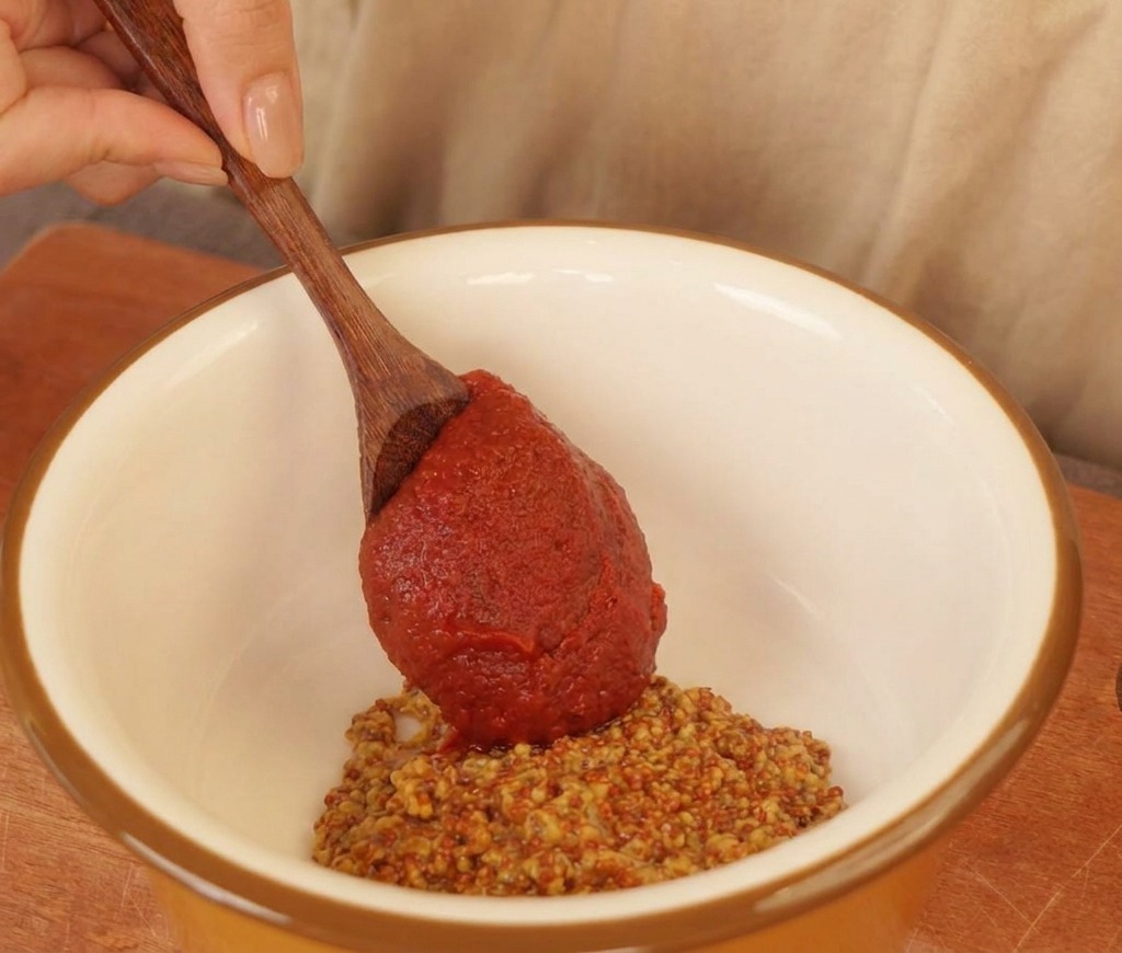 Ein Holzlöffel gibt rote Chilisauce in eine weiße Schüssel mit grobem Senf und Mayonnaise.